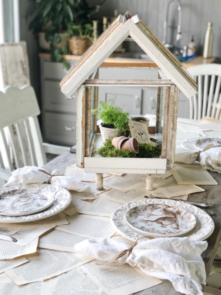 table runner made out of book pages laying across farm table with 4 place settings of white dinner plate layered with white salad plate with brown bunny pattern white linen napkin to the left of plate tied with brown/white striped napkin fork and knife to right of plate small greenhouse for centerpiece with neutral seed packets small terra cotta pots and a small plant inside