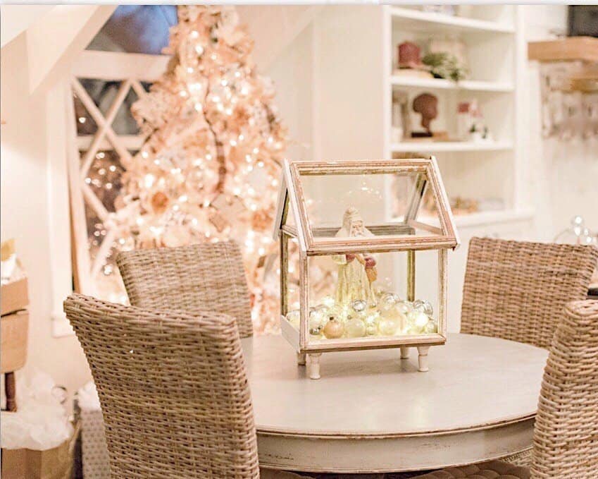 small vintage greenhouse with a white Santa inside and filled with silver vintage Christmas ornaments is the table centerpiece for my round dining room table with 4 woven chairs around it and the white vintage Christmas tree in the background