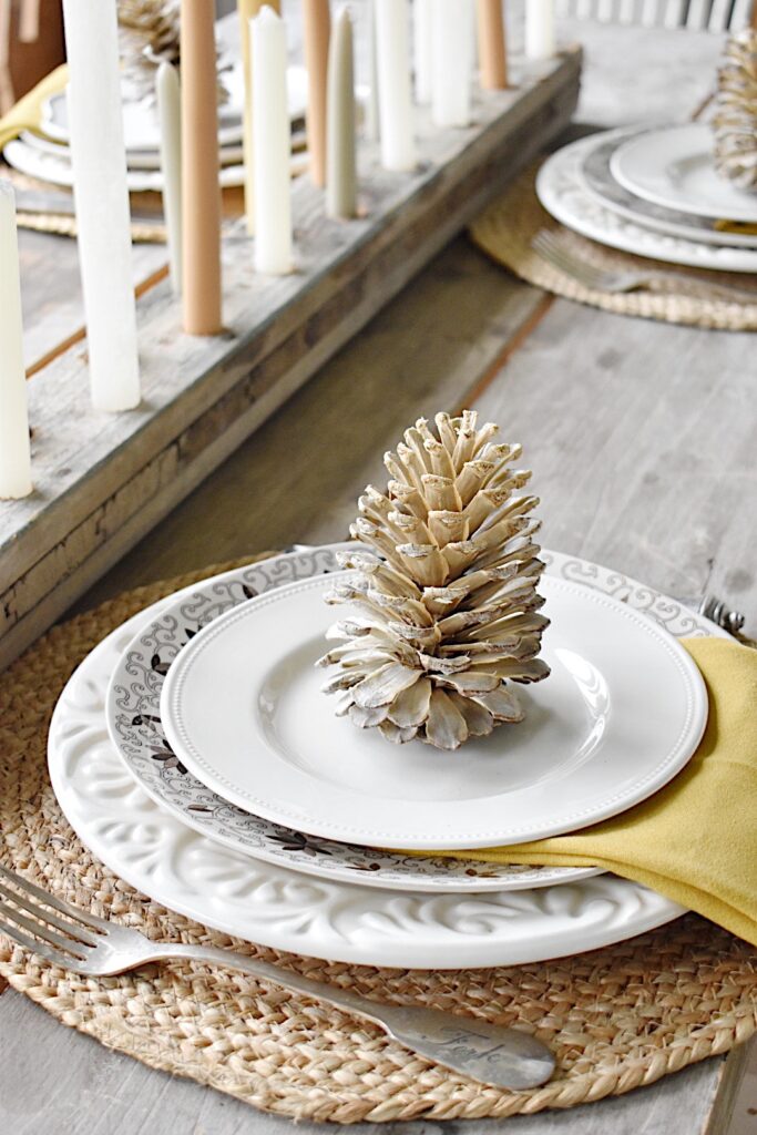 fall tablesetting with bleached pinecone in center of stack of white plates and brown transferware plates and gold napkin between plates 