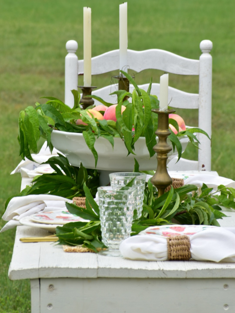 peach themed tablescape set at a long white farmtable. White dinner plates are topped with a peach pattern melamine plate and a white linen napkin with a woven napkin ring sits beside each place setting. The center piece is a large pedestal white bowl filled with fresh peaches and peach tree leaves, and 3 vintage brass candlesticks with white tapers surround the centerpiece.
