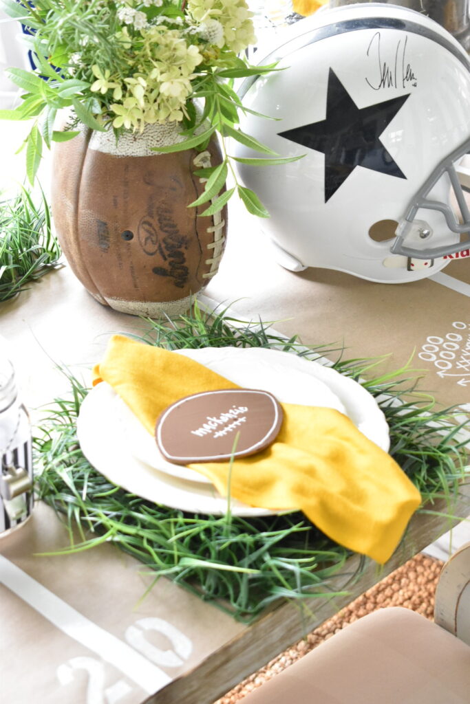 football theme place setting. faux grass square for placemat, white dinner plate stacked with white salad plate, yellow napkin and wooden football cutout on top of napkin for a place card