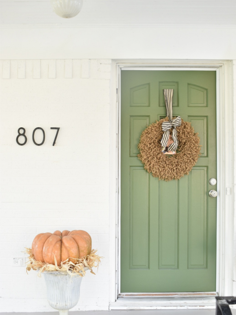front of white cottage with green door white metal urn with large pumpkin on top of urn jute wreath hanging on door