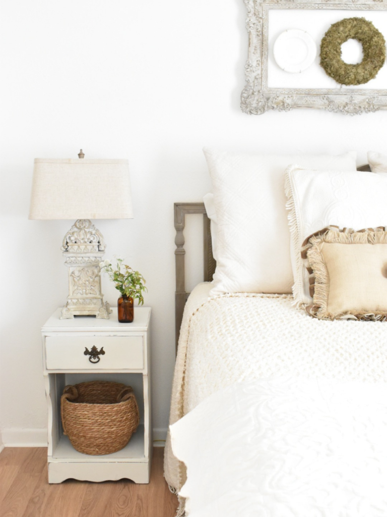 edge of full size bed with cream color bedding small white nightstand with basket on bottom shelf and lamp and vase of flowers on table