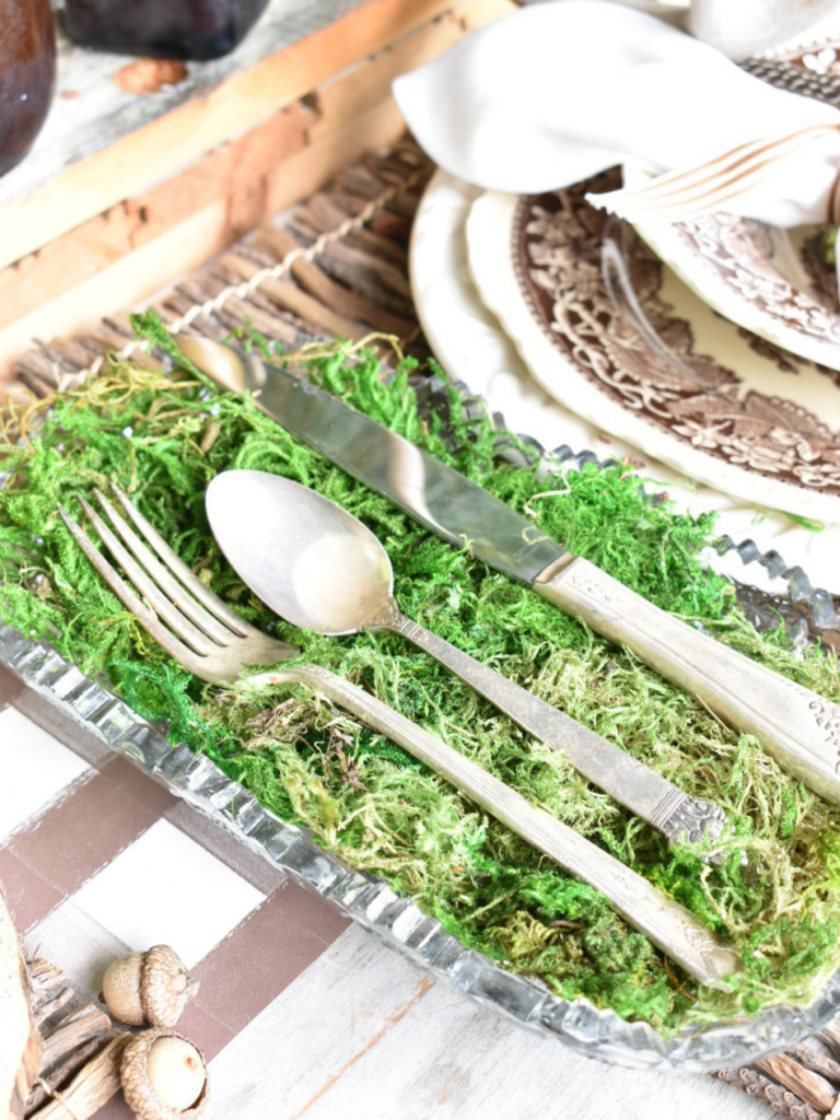 clear glass tray lined with green moss with spoon fork knife laying on top sitting beside place setting