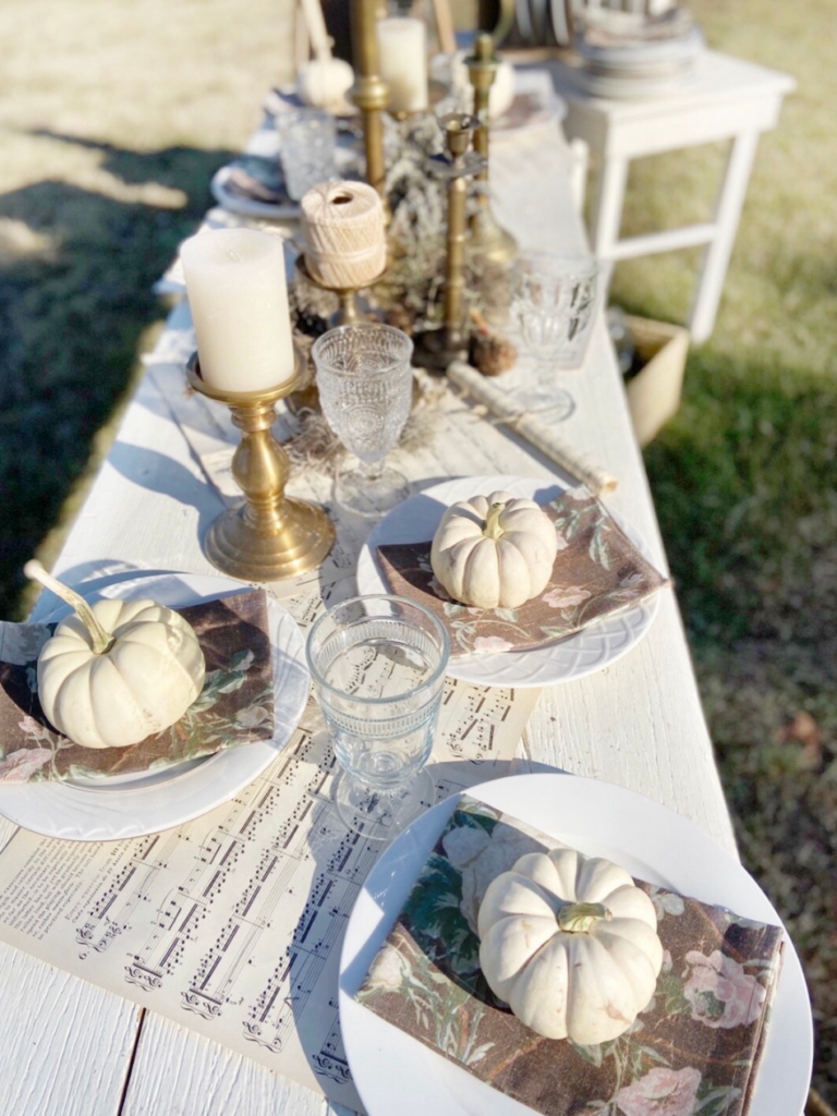 long white farm table sheet music down center as runner white plates with a mini white pumpkin on top of each plate vintage glass goblets at each place and brass candlesticks with white candles down center of table