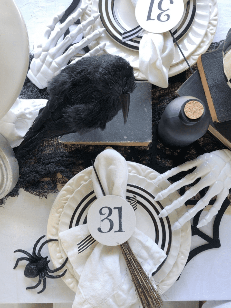 halloween place setting with 2 stacked white plates white salad plate with black stripe border on top of stack white napkin on top of plate with a round card with 31 stamped on it white skeleton hand on edge of plate black felt spider web placemat black crow on top of stack of black books