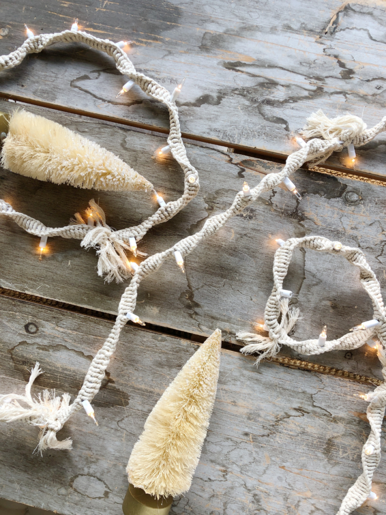 strand of white christmas lights wrapped with macrame laying on a wood table with 2 white botte brush trees beside them