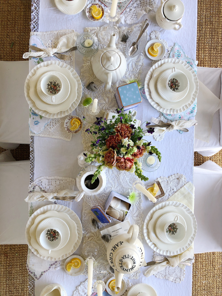 top view of long table covered with white tablecloth and lace table runner 6 place settings with vintage hankie for placemat white dinner plate topped with white fluted edge salad plate then white teacup and saucer silverware to left of plate tied with ivory vintage handkerchief 6 clear glass taper holders with white tapers down center of table white teapot sitting on 2 vintage books with flowers inside and 2 other white teapots one with tea in it 2 tea tins filled with teabags down center of tablesmall white dish beside each place setting with lemon slices for a tea party brunch for girlfriends