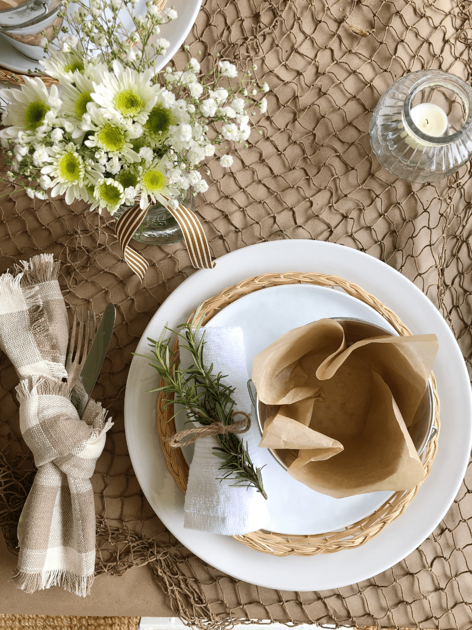 overhead view place setting for shrimp boil table covered in brown paper then fish net and white dinner plate stacked with a woven plate holder then white salad plate on top and small metal bucket lined with parchment paper on top of salad plate fork and knife tied with tan and white check fabric napkin to left of plate and mason jar of daisies to left of plate white washcloth rolled and tied with twine and stem of rosemary tucked in