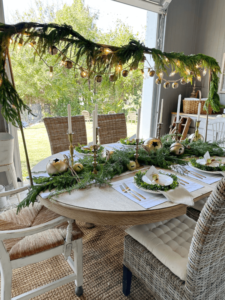 simple christmas tablescape with greenery table runner with large gold bells and brass candlesticks down center of table over the table bar with greenery gold bell garland and white lights overhead and 4 christmas place settings white linen placemat gold flatware white dinner plate small greenery wreath with white salad plate on top gold bell with gold felt ribbon on top of plate for place card holder linen napkin under dinner plate greenery table runner with large gold bells and brass candlesticks