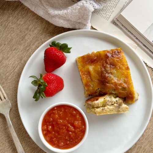 slice brunch egg casserole on white plate with 2 strawberries and small bowl of salsa