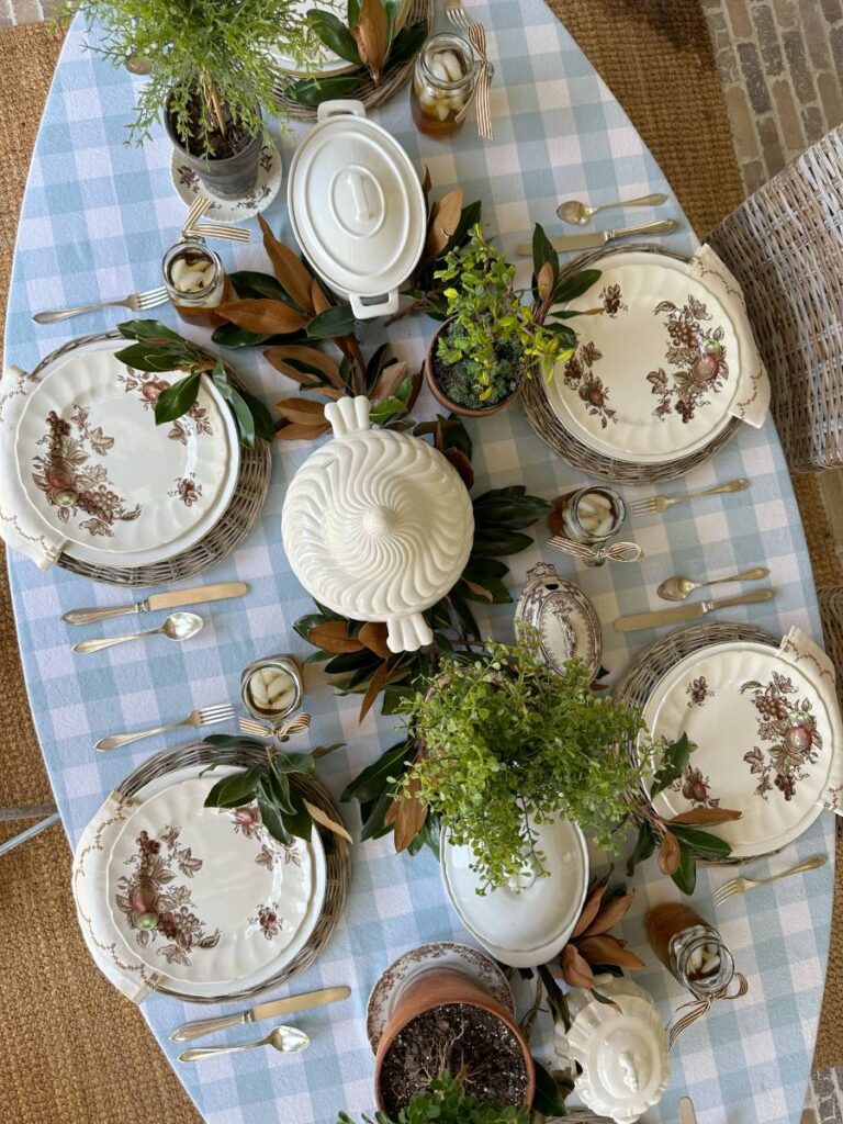 overhead view of tablescape blue check tablecloth transferware plates woven placemats white covered dishes and topiaries and magnolia leaves in center of table for potluck dinner